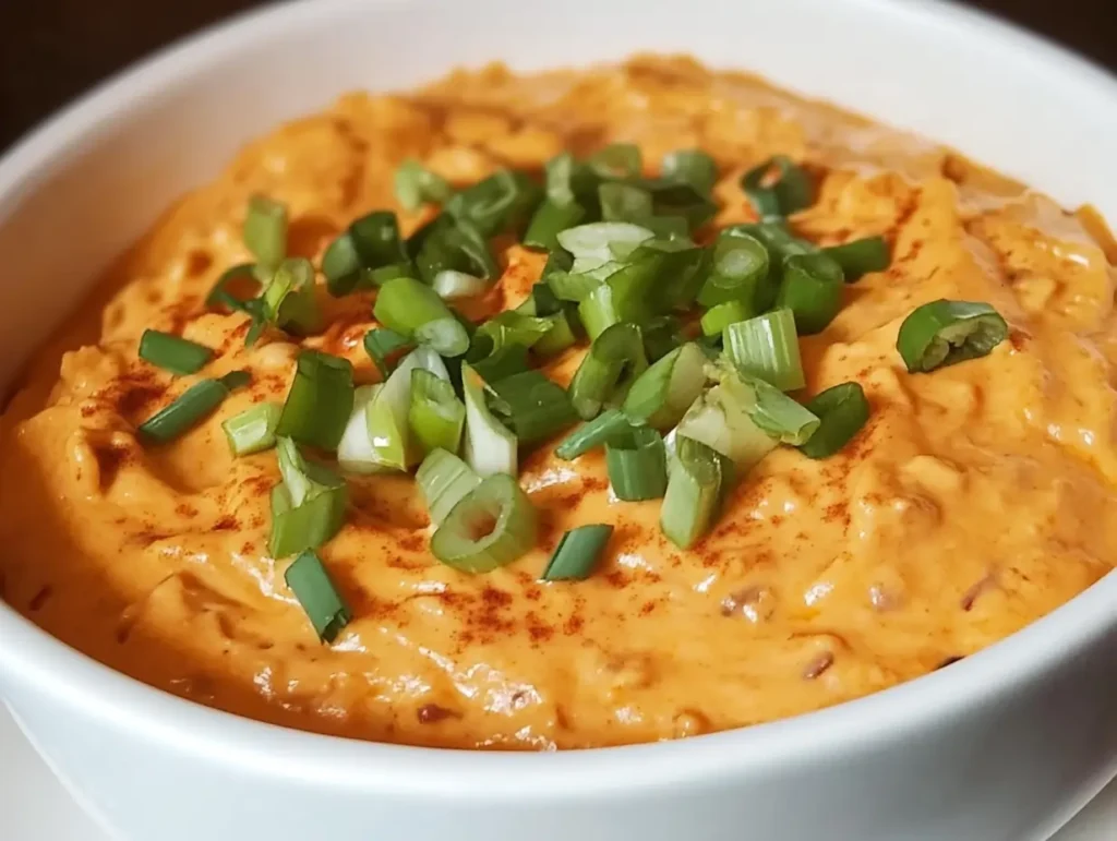 A vibrant bowl of smoked queso dip, creamy and orange, garnished with freshly chopped green onions and a dusting of paprika, served in a sleek white bowl.
