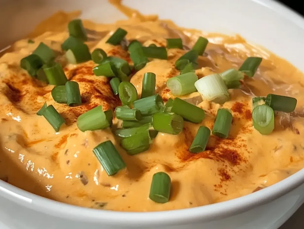 A close-up of creamy smoked queso dip topped with vibrant green onion slices and a sprinkle of paprika, served in a white bowl.