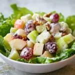 A close-up of a fresh Waldorf salad with apples, grapes, walnuts, and lettuce in a white bowl.