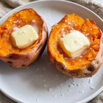 Two perfectly cooked sweet potatoes split open with melted butter and a sprinkle of salt on a white plate.