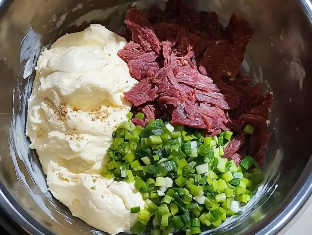 A stainless steel mixing bowl filled with creamy cheese, shredded chipped beef, chopped green onions, and seasonings, ready to be combined for a cheese ball recipe.