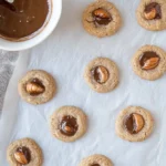 Freshly baked chestnut flour thumbprint cookies filled with melted chocolate and topped with whole almonds, resting on parchment paper with a bowl of chocolate in the background.