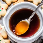 A bowl of sugar cookie syrup with a spoon and festive cookies.