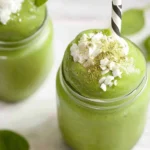 Thick matcha green tea smoothie in mason jars, topped with shredded coconut and matcha powder, with a black-and-white striped straw.